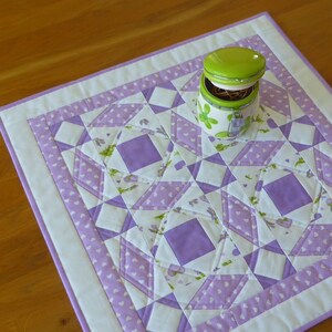 Op de afbeelding: Een paarse en witte gestikte tafelrunner met een groene en witte keramische pot in het midden. De runner heeft een geometrisch patroon met een witte achtergrond en paarse vierkanten.