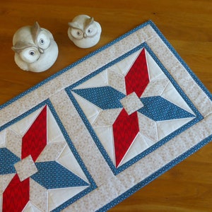Puede incluir: Un camino de mesa con un patrón geométrico con tela roja, blanca y azul. El camino de mesa tiene un fondo blanco con un borde azul y un diseño de estrella roja y blanca en el centro.