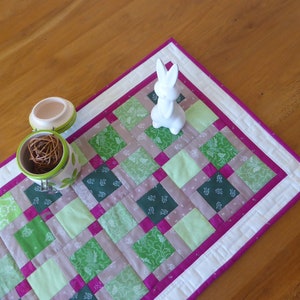 以下が含まれることがあります： 緑、ピンク、白の正方形のパッチワークデザインのキルトテーブルランナー。ランナーは白い縁取りと、縁に沿ってピンクのアクセントが付いています。白いセラミックのウサギの置物がランナーの上に置かれています。