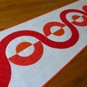 Op de afbeelding: Een witte tafelrunner met een herhalend patroon van rode en oranje cirkels. De cirkels zijn in een golvende lijn in het midden van de runner gerangschikt.