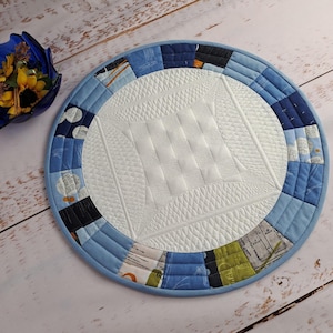 Peut inclure: Un dessous de plat rond en patchwork bleu et blanc avec un centre blanc et une bordure bleue. La bordure est composée de différents tissus bleus avec une variété de motifs.