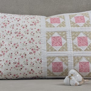 Könnte beinhalten: Ein rechteckiges Zierkissen mit einem Blumenmuster auf der einen Seite und einem Patchwork-Design auf der anderen Seite. Die Blumenseite hat einen weißen Hintergrund mit rosa Blumen. Die Patchwork-Seite hat rosa und beige Quadrate. Vor dem Kissen befindet sich eine weiße Keramikvogel-Figur.