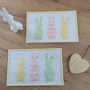 Könnte beinhalten: Zwei rechteckige, gesteppte Tischsets mit gelben Rändern, jeweils mit drei Hasen-Silhouetten in grünen, rosa- und gelbblühenden Mustern. Eine weiße Keramik-Hasenfigur und ein Holzherz sind ebenfalls vorhanden.