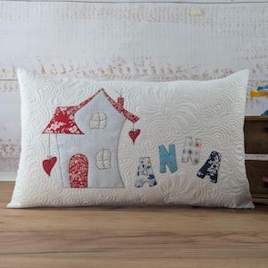 Puede incluir: Cojín decorativo blanco con un patrón acolchado y una aplicación de casa. La casa tiene un techo y una puerta florales rojos, con el nombre "ANNA" aplicado debajo. Un bolso rojo y un dragón de juguete también están presentes.