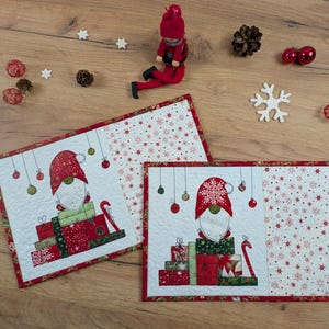 Könnte beinhalten: Zwei rechteckige, gesteppte Platzsets mit einem Wichtel mit roter Mütze, der auf einem Stapel verpackter Geschenke sitzt. Das Design umfasst hängende Ornamente und ein Seitenfeld mit rot-weißem Schneeflockenmuster, eingefasst in rotem Stoff.