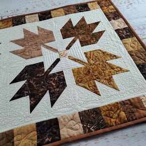 Op de afbeelding: Een gestikte tafelloper in bruin en crème tinten. De loper heeft een centraal ontwerp van vier gestileerde esdoornbladeren, omringd door een rand van kleinere vierkanten in verschillende bruintinten.