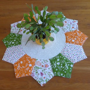 Könnte beinhalten: Ein runder, weißer Tischläufer mit einem Sternmuster. Der Läufer ist aus Stoff mit verschiedenen Blumenmustern in Grün-, Orange- und Weißtönen gefertigt. In der Mitte des Läufers steht eine kleine Topfpflanze.