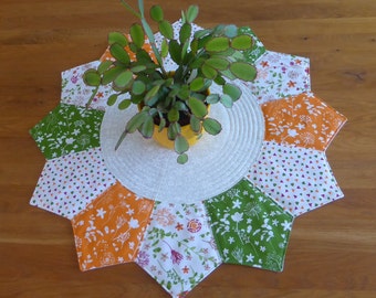 Op de afbeelding: Een ronde, witte tafelrunner met een stervormig ontwerp. De runner is gemaakt van stof met verschillende bloemenpatronen in tinten groen, oranje en wit. Een kleine plant in een pot staat in het midden van de runner.