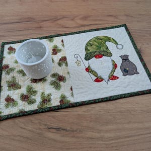 以下が含まれることがあります： クリスマスノームのデザインが施されたキルトプレースマット。ノームは緑色の帽子をかぶり、ランタンを持っています。灰色のバッグがノームの隣にあります。雪の結晶模様の白いキャンドルホルダーがプレースマットの上にあり、松ぼっくりと緑の葉の模様があります。