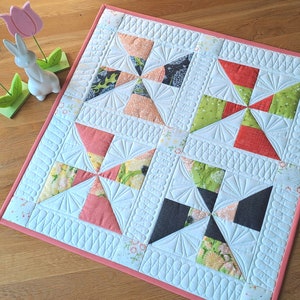 Könnte beinhalten: Eine Decke mit rosa Rand und weißem Hintergrund. Die Decke besteht aus vier Quadraten, jedes mit einem anderen Stoffmuster. Die Muster sind ein grünes und rotes Windrad, ein rosa und gelbes Windrad, ein schwarzes und weißes Windrad und ein grünes und gelbes Windrad.