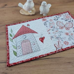 Può includere: Un runner trapuntato con un motivo a casa e farfalla. La casa ha un tetto e una porta rossi, con un cuore appeso. Il runner ha un bordo floreale rosso. Due statuette di uccelli in ceramica bianca sono sullo sfondo.