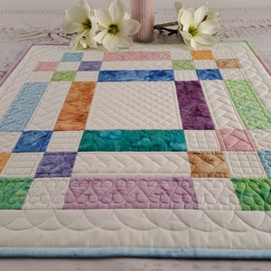 May include: A white quilted table runner with a patchwork design featuring squares of various colors, including orange, green, blue, and purple. The runner has a scalloped edge and is quilted with a variety of patterns.