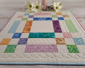 Op de afbeelding: Een witte gestikte tafelrunner met een patchwork-ontwerp met vierkanten in verschillende kleuren, waaronder oranje, groen, blauw en paars. De runner heeft een gegolfde rand en is gestikt met verschillende patronen.
