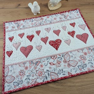 Könnte beinhalten: Weißer, gesteppter Tischläufer mit rotem Rand und Herzdesign. Der Läufer zeigt hängende rote Herzen und ein Blumen- und Schmetterlingsmuster. Dekorative weiße Keramikvögel befinden sich im Hintergrund.