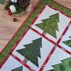 May include: A white quilted table runner with a red and green border. The runner features a repeating pattern of green Christmas trees with different designs.