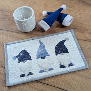 Könnte beinhalten: Ein gesteppter Tischläufer mit drei Zwergenfiguren mit weißen Bärten und blauen Hüten. Der Tischläufer hat einen grauen Rand und liegt auf einer Holzoberfläche, zusammen mit einem Kerzenhalter und zwei kleinen blauen Hüten mit weißem Besatz.