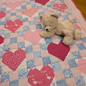 Può includere: Una trapunta patchwork con un motivo ripetuto di quadrati rosa e blu intervallati da grandi applicazioni in tessuto a forma di cuore in varie tonalità di rosa, alcune con pois o motivi di anatre, con un orsacchiotto di peluche beige sopra.