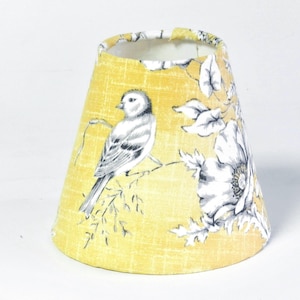 May include: A yellow lampshade with a bird and floral design. The bird is depicted in grey and white, perched on a branch. The flowers are white with grey detailing. The lampshade has a conical shape.