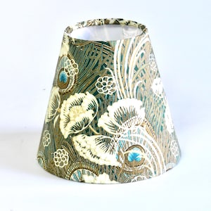 May include: A conical lampshade with a floral design. It features large cream flowers, teal accents, and a dark teal background. The lampshade is lined with white fabric.