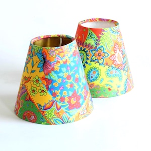 May include: Two lampshades with a patchwork design. The shades feature floral and paisley patterns in yellow, blue, orange, and green. The interior of the lampshades is gold. The lampshades are cone-shaped.