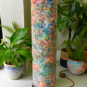 May include: A tall, cylindrical floor lamp with a colorful, abstract design. The lamp's shade features a blend of orange, blue, green, and pink hues. The lamp has a brown cord and sits on a white surface, with potted plants in the background.