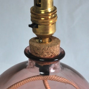 May include: A pink glass bottle lamp base with a cork stopper and brass lamp holder. The lamp has a woven cord and a black on/off switch. The lamp base is a soft pink colour.