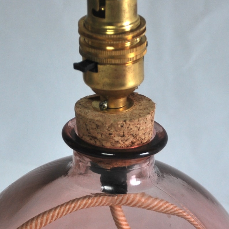 May include: A pink glass bottle lamp base with a cork stopper and brass lamp holder. The lamp has a woven cord and a black on/off switch. The lamp base is a soft pink colour.