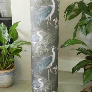 May include: A tall cylindrical floor lamp featuring a dark grey fabric shade decorated with a repeating pattern of white and blue herons amidst green tropical foliage. The lamp stands on small, simple metal feet and includes a power cord.