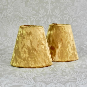 May include: Two conical lamp shades crafted from a golden yellow crushed velvet fabric. The shades feature a smooth, tapered silhouette with a wide base and a narrower top opening, showcasing the textured, light-reflecting surface of the velvet material.