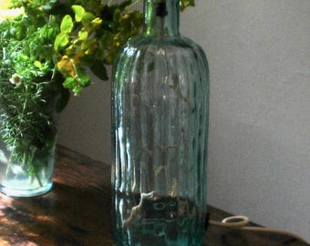 May include: A distinctive table lamp crafted from a textured glass bottle. The lamp has a mustard-yellow lampshade with a floral design. The lamp sits on a wooden surface, with a vase of flowers visible in the background.