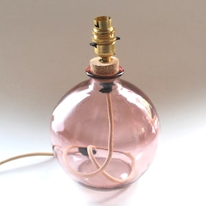 May include: A spherical, pink glass lamp base with a cork stopper and brass bulb holder. A beige cord extends from the base. The lamp is designed to hold a light bulb, suitable for a desk or bedside table.