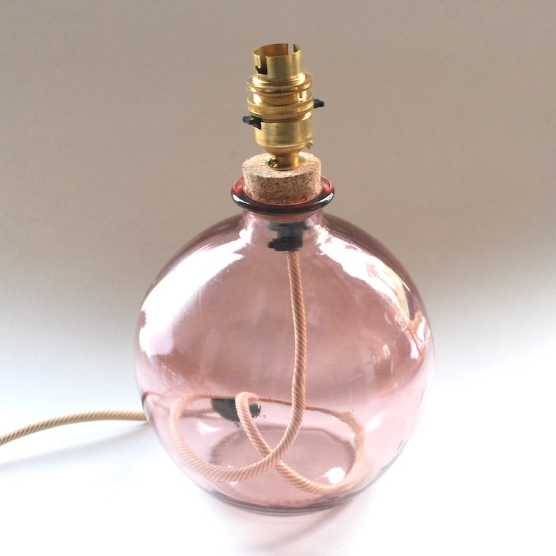 May include: A spherical, pink glass lamp base with a cork stopper and brass bulb holder. A beige cord extends from the base. The lamp is designed to hold a light bulb, suitable for a desk or bedside table.