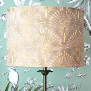 May include: A cylindrical lampshade in beige, adorned with a gold, fan-shaped design. It is mounted on a dark metal lamp base. The backdrop is a teal wallpaper with a floral and bird pattern.