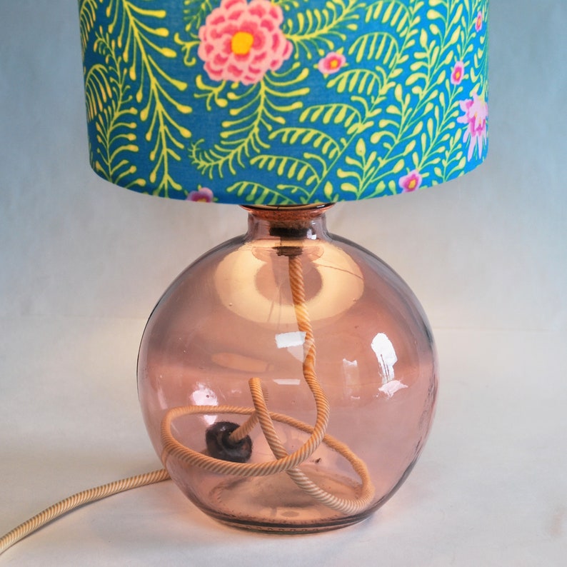 May include: A table lamp with a spherical, pink glass base and a patterned lampshade. The lampshade has a blue background with yellow and pink floral patterns. The lamp features a rope-style cord and a cork stopper.