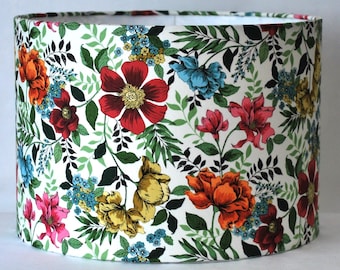 May include: A cylindrical lampshade featuring a white background with a dense, colourful botanical pattern. The design includes large red, orange, yellow, and pink flowers interspersed with small blue blossoms and various green leafy foliage.