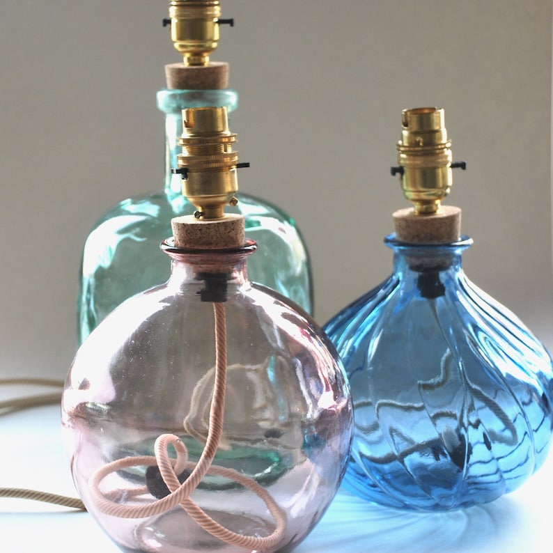 May include: Three decorative glass bottle lamps in shades of blue, green, and pink. Each lamp has a cork stopper, brass bulb holder, and a fabric-covered cord. The lamps are designed with a vintage aesthetic.