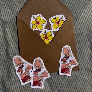 Könnte beinhalten: Ein brauner Umschlag mit drei gelben Hibiskusblüten-Aufklebern und drei Aufklebern einer Person in einem weißen Crop-Top und rotem Rock. Die Aufkleber befinden sich auf einem gestreiften grauen Hintergrund.