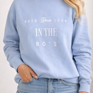 Op de afbeelding: Een lichtblauw sweatshirt met de witte tekst "ESTD Born 1986 IN THE 80'S". Het sweatshirt heeft een ronde hals en wordt gecombineerd met een blauwe jeans. De stijl is casual en heeft een retro uitstraling.