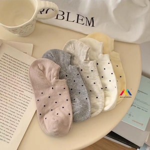 Könnte beinhalten: Eine Auswahl an Füßlingen in verschiedenen neutralen Farben, darunter Beige, Grau und Creme, jeweils mit kleinen schwarzen Polka-Dots verziert. Die Socken sind auf einer hellen Oberfläche angeordnet, was auf ein lässiges Thema für den Alltag hindeutet.