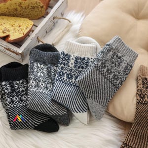 Könnte beinhalten: Eine Auswahl an Wintersocken mit Schneeflocken- und Streifenmustern in Grau-, Schwarz- und Cremetönen. Die Socken sind auf einer flauschigen, weißen Oberfläche angeordnet. Im Hintergrund ist ein Brotlaib zu sehen.