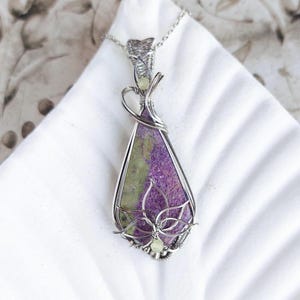 May include: A handcrafted pendant necklace featuring a teardrop-shaped stone with purple and green hues, encased in a silver wire frame. The pendant has a floral design at the bottom and hangs from a delicate silver chain.