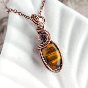 Könnte beinhalten: Eine kupferfarbene Anhängerkette mit einem Tigeraugenstein. Der ovale Stein ist in einem drahtumwickelten Design eingefasst. Die Kette ist ebenfalls kupferfarben, und der Anhänger wird auf einer weißen Oberfläche präsentiert.