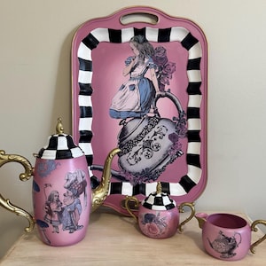 May include: A pink and white tea set with an Alice in Wonderland theme. The teapot, creamer, and sugar bowl feature black and white checkered lids and Alice in Wonderland illustrations. The tray is pink with a black and white checkered border and features an illustration of Alice falling down the rabbit hole.