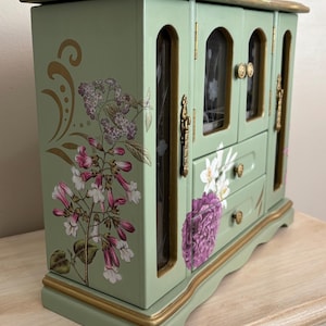May include: A sage green jewelry box with gold trim and floral decals. The box features two glass-fronted doors, two drawers, and gold hardware. Floral designs include white, pink, and purple flowers. The box sits on a light-colored surface.