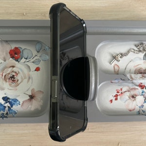 May include: A gray tray with four compartments, each with a floral design. The tray has a phone holder, a ring holder, and two compartments for jewelry. The phone holder is black and has a pop socket attached to the back of the phone.