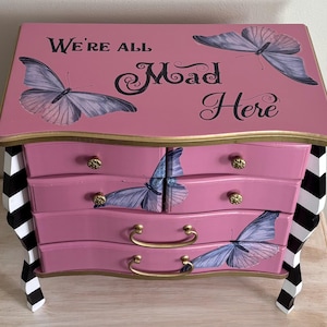 May include: A pink wooden chest of drawers with four drawers, featuring purple butterfly illustrations and the text "We're all Mad Here" on the top surface. The piece has gold-toned hardware and four legs painted with a black and white striped pattern.