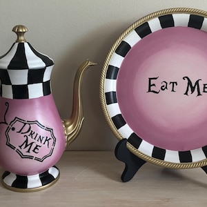 May include: A pink and white teapot with a black and white checkered pattern and the words "Drink Me" on the side, and a pink and white plate with a black and white checkered pattern and the words "Eat Me" on the center.