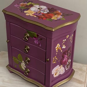 May include: A plum-colored jewelry box featuring four drawers with brass-toned pulls and gold-painted edges. The exterior is decorated with floral decals in shades of red, purple, white, and green on the top surface and the side panel.