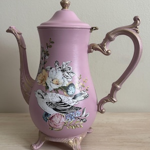 May include: A pink decorative teapot with a bird and floral design. The teapot has a gold-colored handle, spout, and base. The lid is topped with a gold finial. The teapot is decorated with a bird, flowers, and leaves.