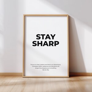 Puede incluir: Un cartel enmarcado con las palabras "STAY SHARP" en letras negras en negrita sobre un fondo blanco. El cartel está en un marco de madera marrón claro y está apoyado contra una pared blanca sobre un suelo de madera. El texto de abajo dice: Concéntrate en lo que importa y bloquea las distracciones.